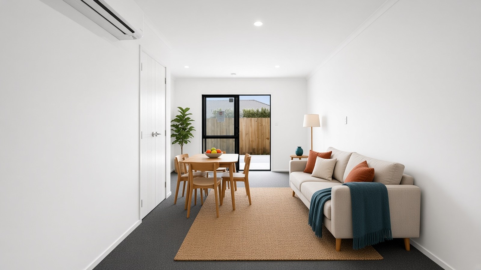Long living room with a heat pump, a wooden dining table with a fruit bowl, and a cream sofa with a teal throw on a jute rug, leading to a glass door.